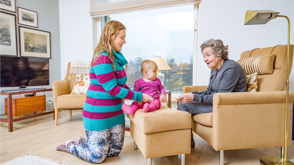 PPå bilden sitter en gammal kvinna i en fåtölj med en kvinna och ett litet barn som sitter på en liten fåtölj.