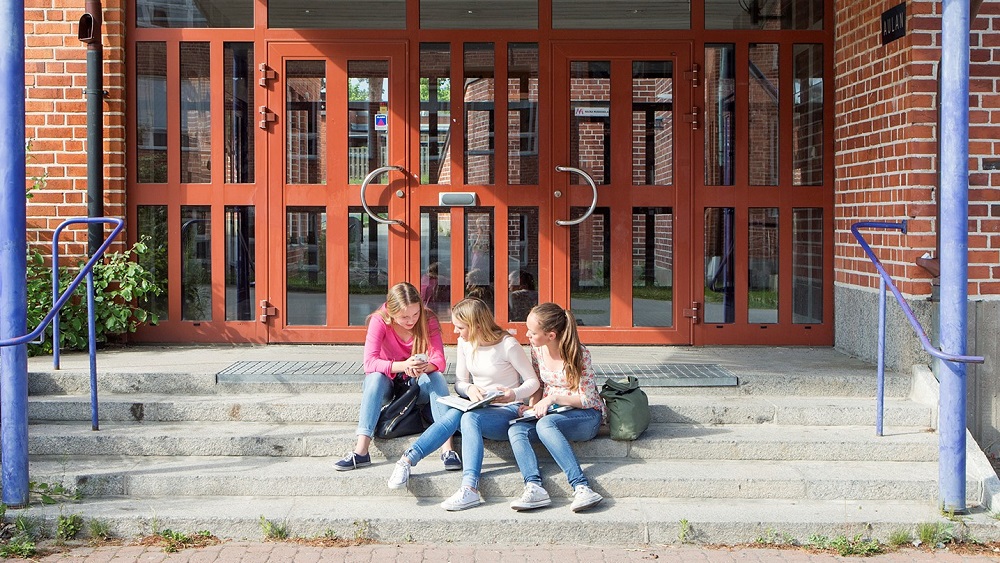 Kolme oppilasta istuu portailla koulun oven edessä.